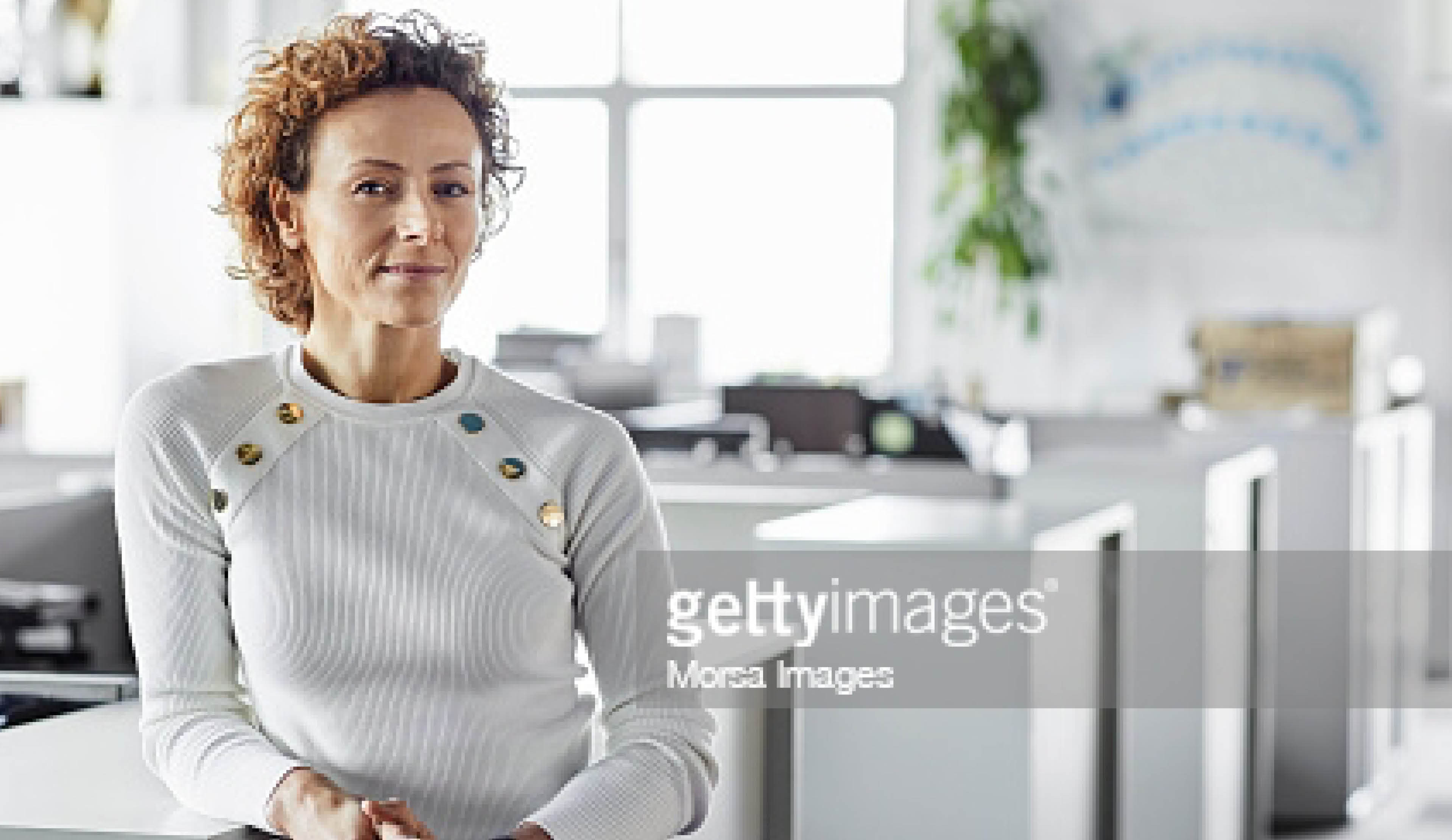 Portrait of confident mature female entrepreneur at workplace  Blond Professional is leaning on cabinet at office  Businesswoman is standing with hands clasped 